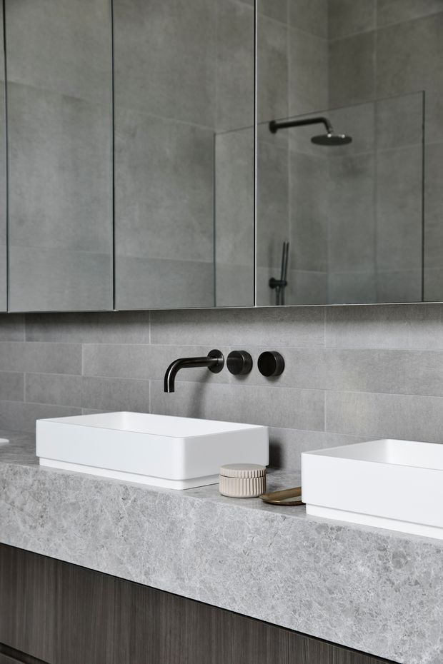 Contemporary double bathroom vanity with two white rectangular vessel basins set on a thick grey stone benchtop. The wall is finished in large-format grey tiles, with matte black wall-mounted tapware and a matching shower visible in the mirror reflection. Mirrored wall cabinetry provides storage above the vanity, while dark timber-toned cabinetry sits below. The composition is clean, restrained and monochromatic with subtle warm accents.