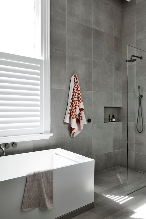 A bright contemporary bathroom featuring a white freestanding rectangular bathtub beside a large white louvered window, grey large-format tiled walls and floor, and a frameless glass walk-in shower with dark fixtures and a recessed wall niche. Neutral grey and white finishes are accented by a patterned rust-red and white towel.