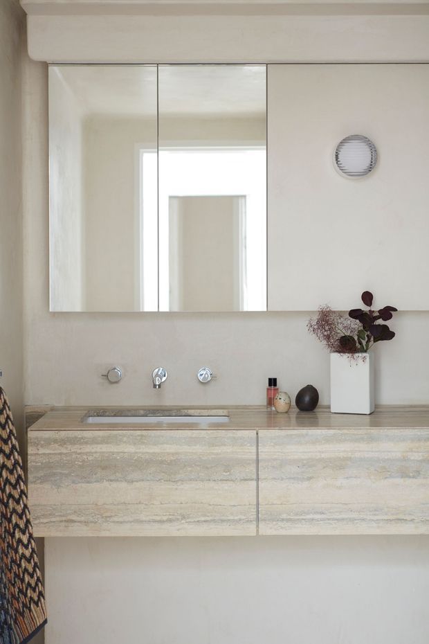 A refined contemporary bathroom vanity with a wall-mounted double cabinet in pale beige stone, an integrated basin, wall-mounted chrome tapware, and a large mirrored cabinet above. The room has warm neutral plastered walls, a small round wall light, and restrained decorative styling with a white vase and dark botanical arrangement.