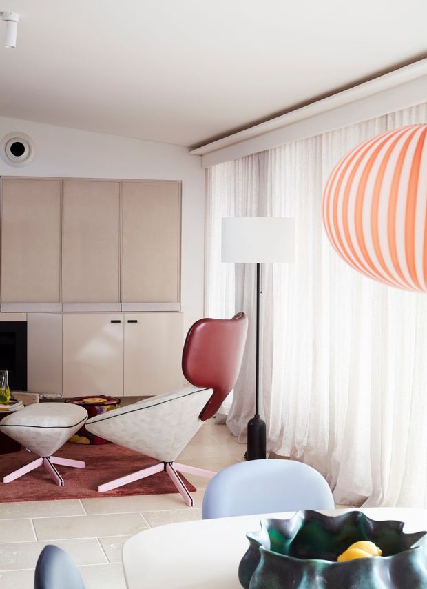 A bright, refined mid-century-inspired living and dining area with a soft neutral palette. Built-in pale cabinetry lines the rear wall, while a sculptural lounge chair with a red leather headrest, a white tulip-style chair, and a rounded pale-blue chair provide curved forms and restrained colour accents. Sheer full-height curtains diffuse daylight beside a black floor lamp, and a large orange-and-white striped pendant adds a bold playful focal point. Pale tiled flooring and a muted rose rug complete the space.