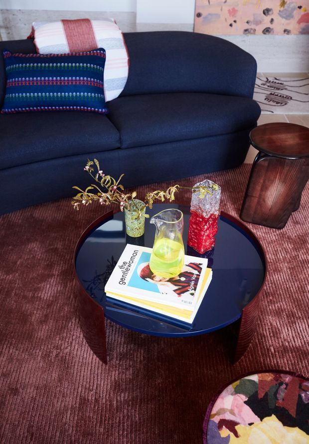 A styled living room vignette dominated by a deep navy upholstered sofa with colourful patterned cushions. A glossy dark round coffee table holds books, a glass pitcher and decorative vessels with branches. The room features a ribbed burgundy-toned carpet, a small timber side table, pale textured wall surfaces and colourful abstract artwork. The overall look is eclectic, colourful and retro-inspired.