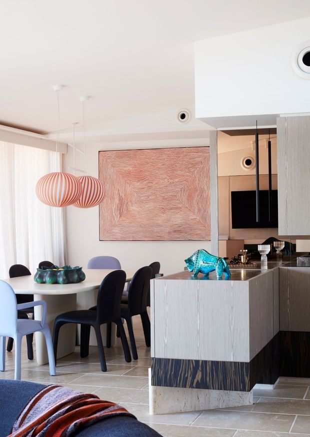 A bright open-plan interior combining a dining area and kitchen, viewed from a living room. The dining space has a round white table, dark upholstered chairs, light blue seating, sheer curtains, and two striped pendant lights. The kitchen features pale vertically textured cabinetry, a dark timber-look island base, stone benchtops, integrated appliances, and black pendant lighting. A large pink-toned textured artwork and colourful sculptural decor add visual interest.