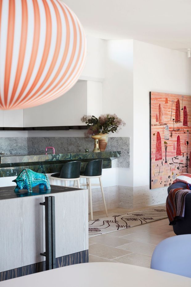 Bright open-plan interior showing a contemporary kitchen and adjacent living area. The kitchen features pale textured cabinetry, a dark stone island and benchtop, a speckled stone splashback, black bar stools, and a vivid pink tap. A large sculptural striped pendant dominates the upper left, while colourful artwork, patterned flooring, and decorative objects add playful character. The edge of a sofa and a dining or foreground table are visible, connecting the kitchen to the living space.