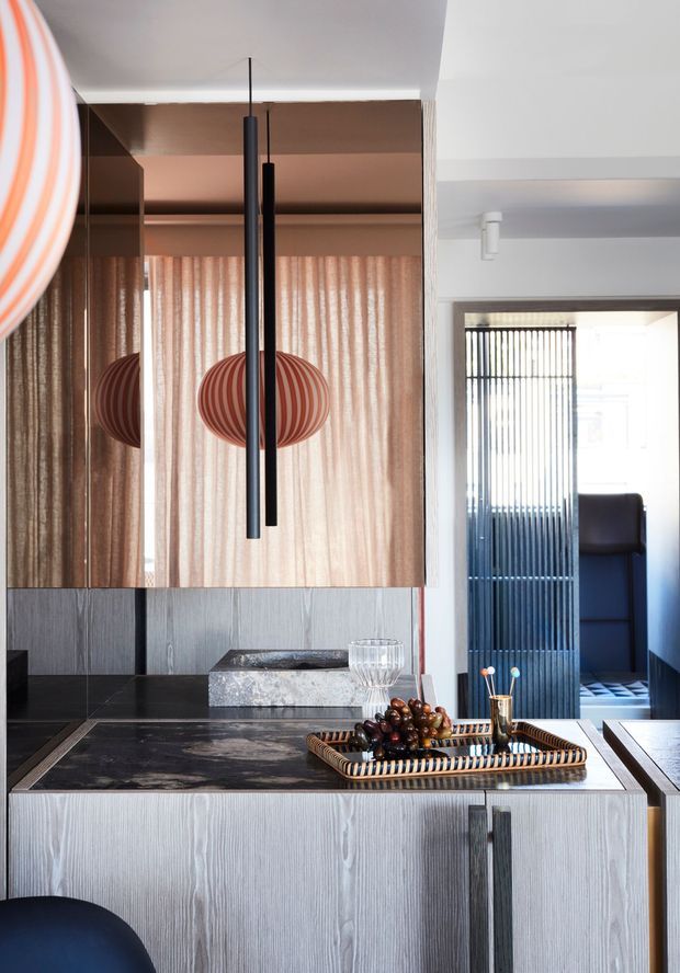 A refined contemporary kitchen or entertaining area with a dark stone island benchtop, vertically grained light-grey timber cabinetry, mirrored upper cabinetry, and warm sheer curtains. Two slim black pendant lights hang above the island, with sculptural red-orange striped pendant shades reflected in the mirrors. A tray with fruit and glassware sits on the island, and a slatted doorway opens toward an adjacent space.