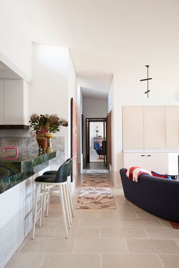 Bright, narrow open-plan interior combining a compact kitchen and living area with a central hallway leading to a home office or study. The space has white walls and cabinetry, a patterned stone backsplash and dark green stone island benchtop, pale large-format tiled flooring, contemporary black bar stools, a dark sofa, and a sculptural black pendant light. A runner and artwork add visual interest, while the high ceiling and long sightline make the room feel airy and elongated.