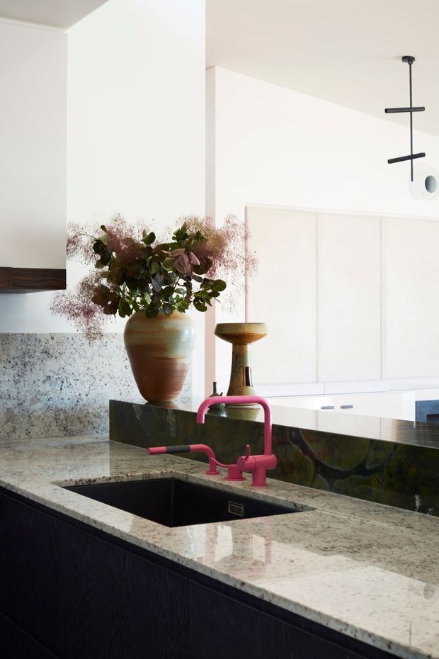 A refined contemporary kitchen focused on a dark island with an integrated rectangular sink and distinctive pink-red mixer tap. The island has a pale, heavily veined stone benchtop and dark textured cabinetry, with a dark reflective splashback behind it. White paneled cabinetry and walls create a bright backdrop, while a ceramic vase with foliage and a sculptural pendant light add decorative detail.