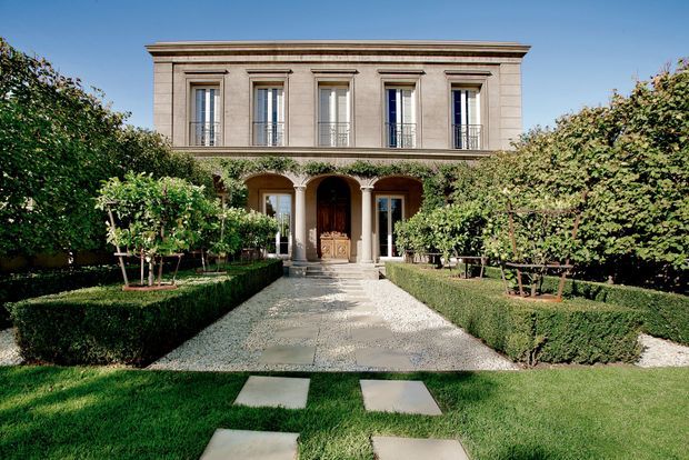 Formal front elevation of a symmetrical two-storey residence set behind a manicured garden. The rendered or stone-toned facade features classical detailing, a strong cornice, tall narrow upper-level windows with wrought-style railings, and a columned entrance with an arched doorway. A gravel and stone path passes through clipped hedges and neatly shaped trees toward the entry.