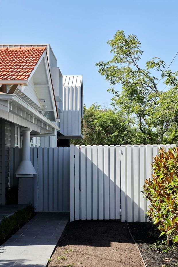 Exterior view of a renovated residential property in bright daylight. A white and pale-grey timber-clad building combines a traditional section with a red terracotta tiled pitched roof and a newer projecting volume with vertically ribbed metal cladding. A tall white picket-style boundary fence, paved walkway, dark garden beds, and established leafy planting occupy the foreground.