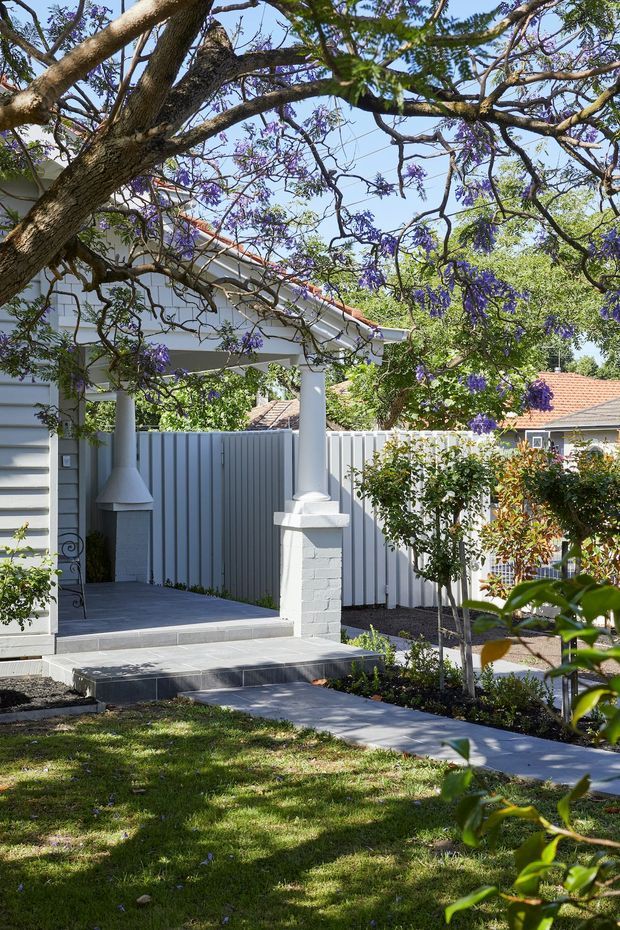 A landscaped front garden and entry porch of a light-coloured residential home, framed by a large flowering tree with purple blossoms. The porch has white columns, a pitched tiled roof with terracotta edging, a grey paved floor and steps, a white-painted brick pillar, and a pale vertical-panel privacy fence. Neatly clipped shrubs and a lawn lead to the covered entrance, with neighbouring tiled-roof houses visible in the background.
