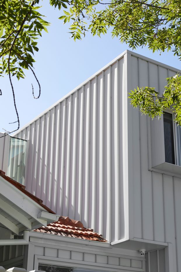 Exterior architectural photograph of a contemporary white-clad building extension rising above an older structure. The facade features repeated narrow vertical ribs or battens, crisp geometric edges, and a projecting dark-framed window. A lower section has traditional white trim and a weathered terracotta tiled roof, creating a clear contrast between the modern upper volume and the existing building. Tree branches frame the image against a bright blue sky.