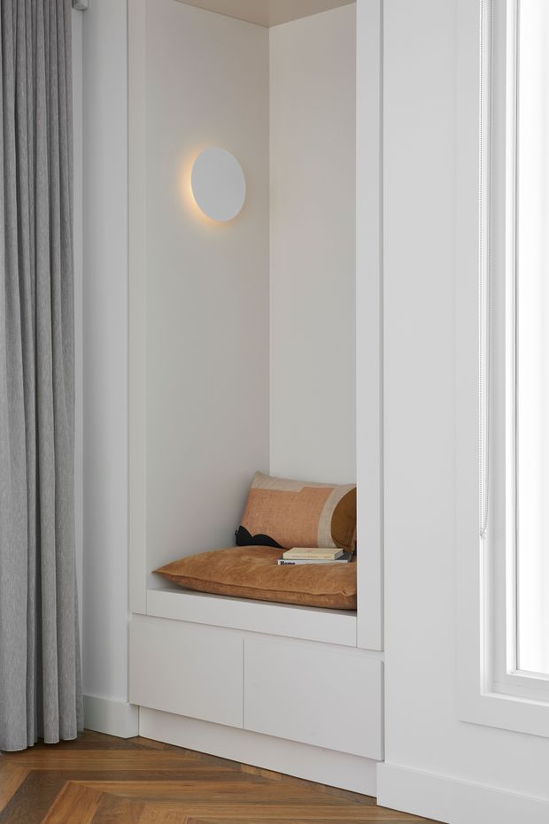 A compact built-in window-seat nook with white painted joinery, a cushioned bench, warm tan pillows, and an integrated round wall sconce. Full-height grey curtains frame the left side, while a bright window is visible on the right. The room has a warm herringbone timber floor and a calm, minimalist contemporary appearance.