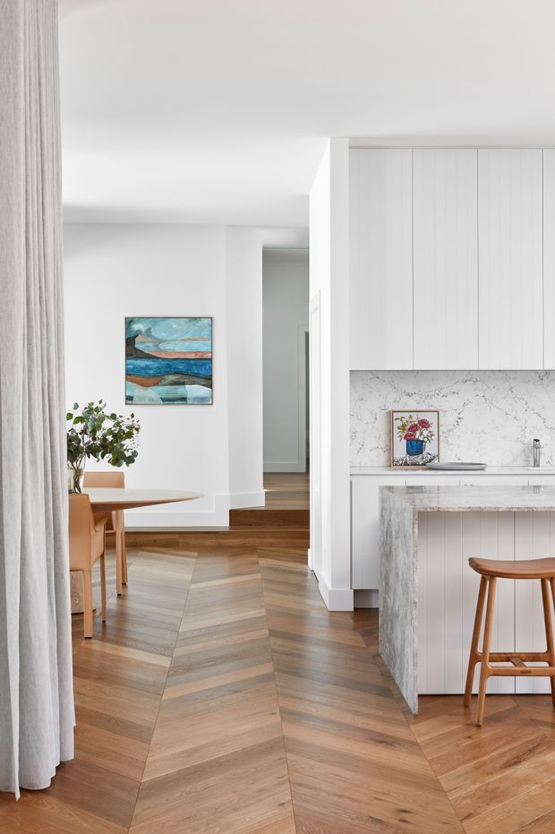 Bright, refined open-plan kitchen and dining interior with white vertical-panel cabinetry, a marble-look splashback and island, warm timber herringbone flooring, and a small round timber dining table. A hallway and stair area extend beyond the kitchen, with contemporary abstract artwork and soft white curtains adding restrained colour and texture.