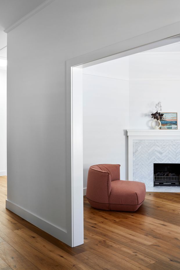 Bright, refined interior view through a wide cased opening into a living room. The room features white walls and detailed cornice mouldings, warm timber flooring, a sculptural rust-coloured lounge chair, and a white fireplace with a pale herringbone tile surround and simple mantel. A small artwork and vase sit above the fireplace.