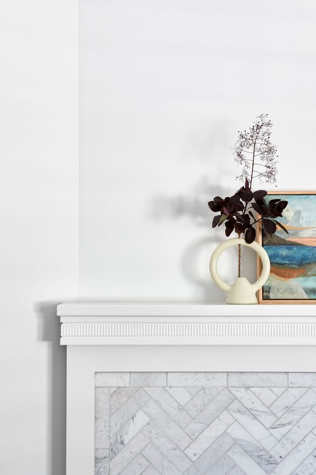Close-up interior detail of a classic white fireplace mantel with layered decorative moulding and a herringbone-patterned marble fire surround. A sculptural cream ceramic vase with dark foliage and a partially visible abstract artwork sit on the mantel against a white wall.