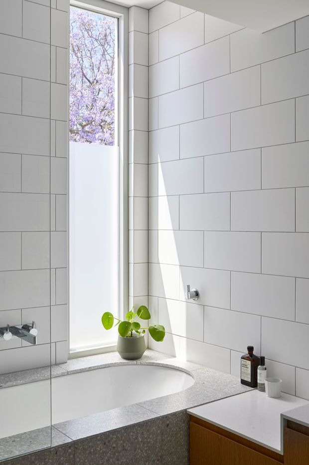 Bright contemporary bathroom with large white rectangular wall tiles, a freestanding oval bathtub set beneath a tall narrow window, and a grey terrazzo-style tiled bath surround and vanity benchtop. A mirrored wall reflects the tub area, while timber-toned vanity cabinetry and minimal toiletries add warmth. Natural light creates a sharp diagonal highlight across the tiled wall.