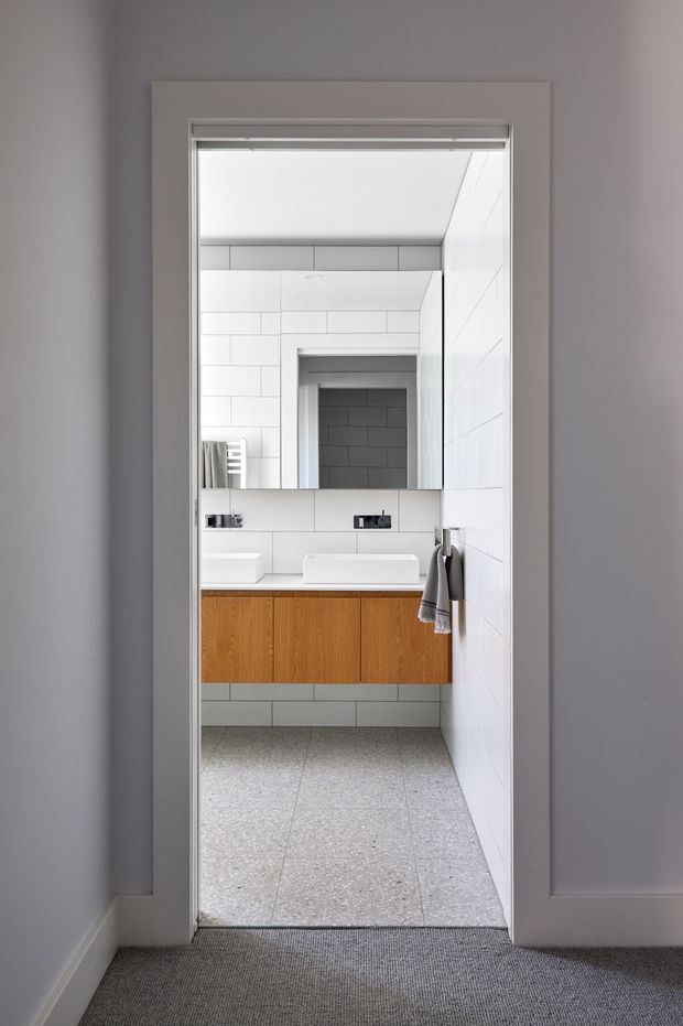 A compact contemporary bathroom viewed through an open doorway, featuring white tiled walls, a large mirror, wall-mounted black tapware, two white vessel basins, and a floating timber vanity with warm wood cabinetry. The bathroom has a pale terrazzo-style tiled floor, grey painted surrounding walls, and a neutral grey carpeted hallway outside.