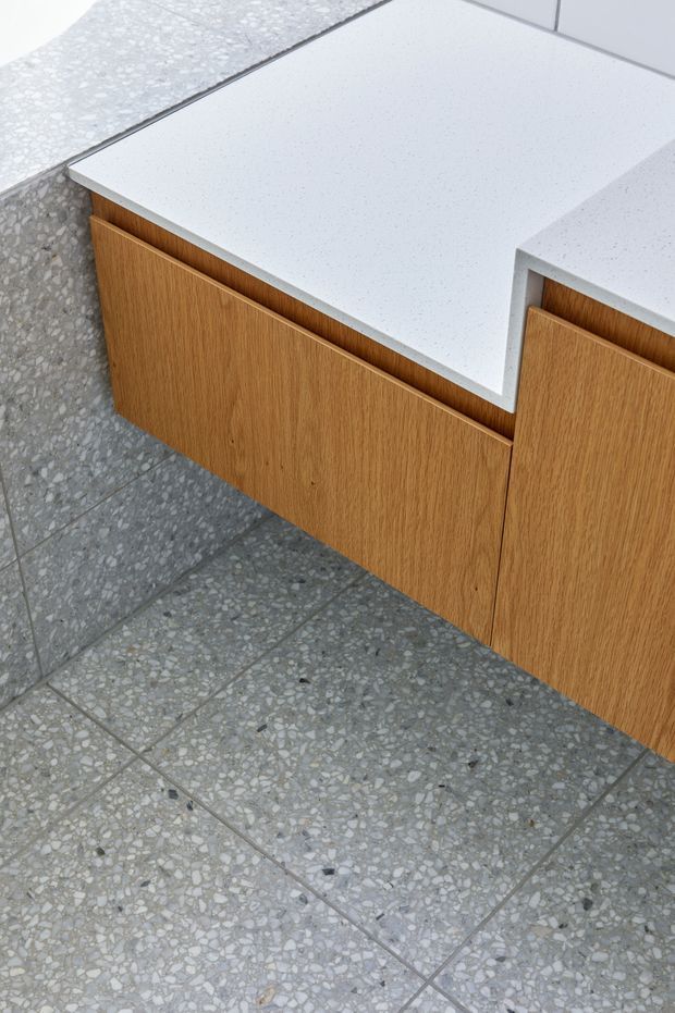 Close-up detail of a contemporary built-in timber vanity or cabinet with warm oak-toned flat-panel fronts and a white speckled engineered-stone benchtop. The cabinetry sits against a grey terrazzo-tile floor and matching terrazzo surface, highlighting clean geometric joinery and contrasting materials.