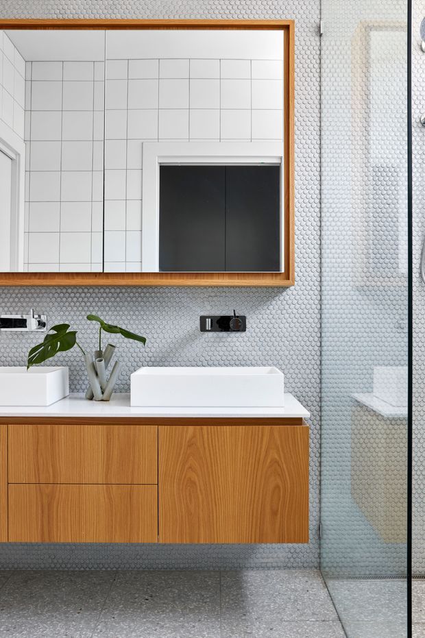 A contemporary bathroom vanity with a floating warm timber cabinet, white countertop and rectangular vessel basin. The walls and shower enclosure are clad in small round grey mosaic tiles, with a timber-framed mirror, black wall-mounted tapware and a clear glass shower screen. Large grey terrazzo-style floor tiles complete the clean, modern composition.
