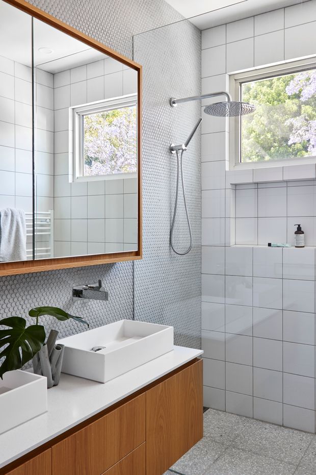 Bright contemporary bathroom featuring a white vessel basin on a timber-veneer vanity, large timber-framed mirror, white square wall tiles, a textured small-format tiled shower wall, chrome fixtures, a recessed shower shelf, and terrazzo-style flooring. Natural light enters through two windows overlooking leafy vegetation.