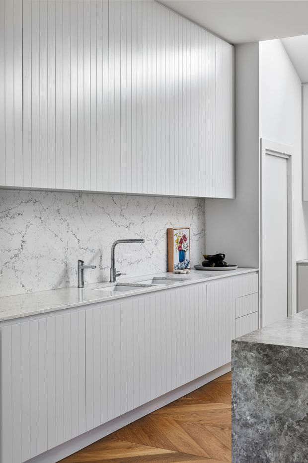 A bright, minimalist kitchen with white vertically fluted cabinetry, a pale veined stone splashback and benchtop, stainless-steel fixtures, and a contrasting dark grey stone island in the foreground. Warm herringbone timber flooring adds texture beneath the predominantly monochromatic scheme.