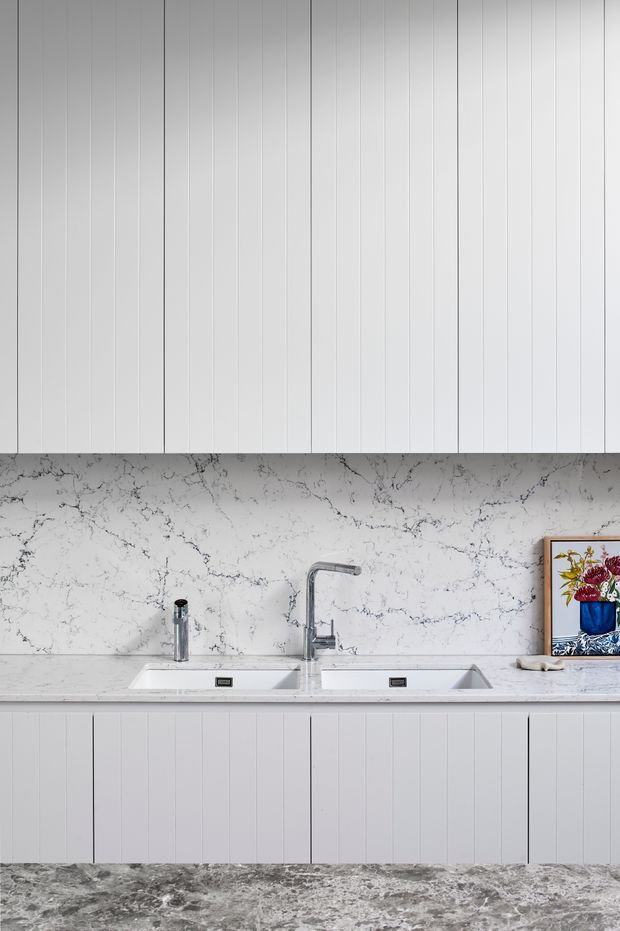 A bright, minimalist kitchen or scullery wall featuring white vertically grooved cabinetry above and below a double-basin undermount sink. The work area has a polished grey-and-white veined stone splashback and benchtop, a chrome gooseneck tap, a separate soap dispenser, and grey stone-look flooring. A framed floral artwork adds a small decorative accent at the right.
