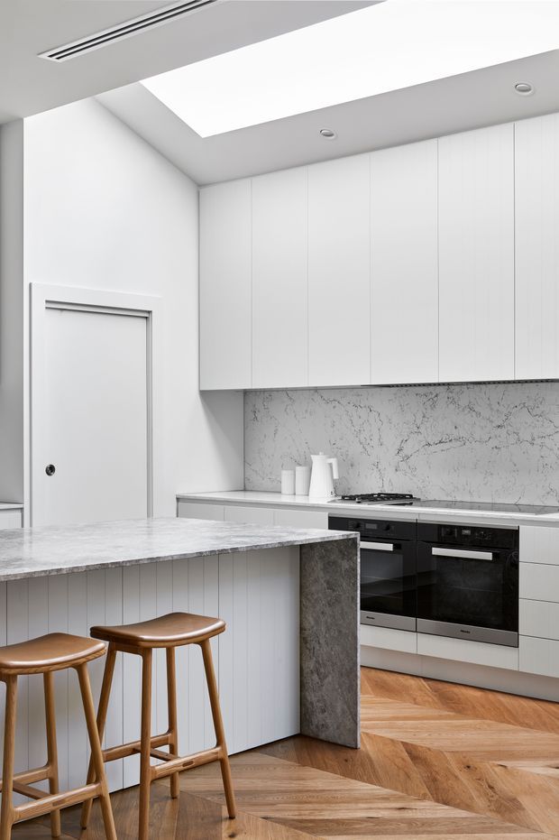 A bright, refined contemporary kitchen with extensive white flat-panel cabinetry, a veined marble-look splashback and benchtops, integrated black appliances, and a large island with sculpted stone sides. Timber bar stools and warm herringbone timber flooring add contrast. A large ceiling skylight and recessed linear ventilation create an airy, minimalist feel.