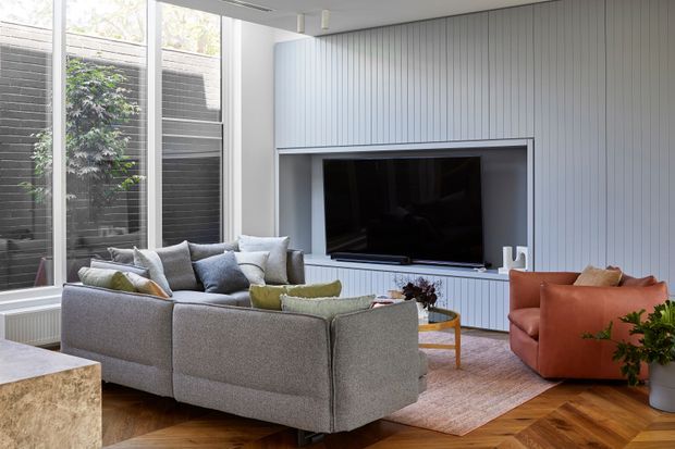 Bright contemporary living room with a large grey sectional sofa arranged around a wall-mounted television. The room features white vertical wall panelling, tall floor-to-ceiling glazing, a warm herringbone timber floor, a pale area rug, a rust-orange lounge chair, and a small timber side table. The space feels airy, refined, and inviting.