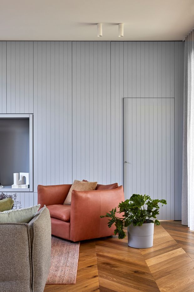 A refined living room corner with a warm terracotta leather armchair, textured neutral sofa, patterned rug, potted plant, and herringbone timber flooring. The room features a full-height vertically panelled grey wall, concealed panel door, recessed shelving, sheer curtains, and simple white ceiling spotlights. The palette balances cool grey architectural finishes with warm timber and leather furnishings.
