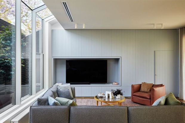Bright contemporary living room with a large grey sectional sofa arranged around a round coffee table, a rust-coloured leather armchair, and a wall-mounted television set within built-in cabinetry. The room features full-height corner glazing, pale vertical wall panelling, recessed linear air conditioning, and a clean neutral palette.