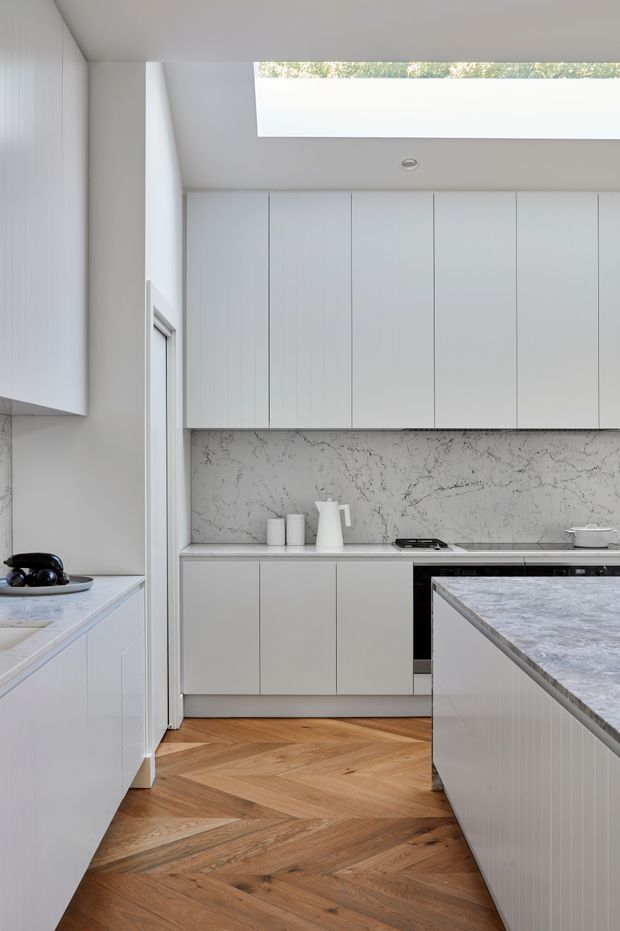 A bright, refined contemporary kitchen with full-height white vertically grooved cabinetry, a veined pale stone backsplash, and a large matching island. Warm timber herringbone flooring contrasts with the cool white and grey surfaces. A long horizontal skylight provides natural light, while minimal white ceramic accessories and integrated appliances maintain a clean, streamlined appearance.