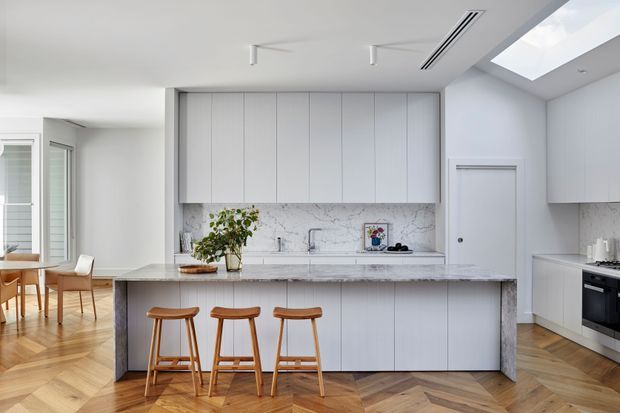 Bright contemporary kitchen with a large central island, three timber stools, pale vertical-panel cabinetry, a marble-look splashback and benchtop, and integrated appliances. The kitchen opens to a dining area with timber herringbone flooring and receives additional daylight through a large skylight.