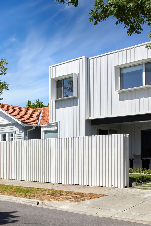 Street-level exterior view of a contemporary two-storey home addition with crisp white vertical-panel cladding, deep-set rectangular windows, and a sheltered ground-floor entry or outdoor sitting area. A tall matching white privacy fence fronts the property. The modern extension contrasts with an older neighbouring or attached weatherboard cottage featuring a terracotta tiled roof. Suburban streetscape with concrete footpath and driveway under a bright blue sky.