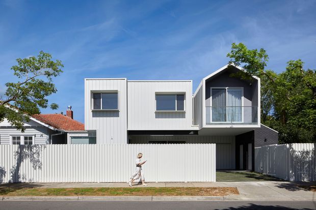 Street-facing view of a contemporary multi-level residence with a white vertically clad box-like addition, projecting upper volume, and a prominent gabled glazed bedroom or living space. The house is screened by a tall white vertical-slat fence, with a narrow driveway and covered entry beneath the upper floor. A neighbouring weatherboard house and mature trees are visible at the sides.
