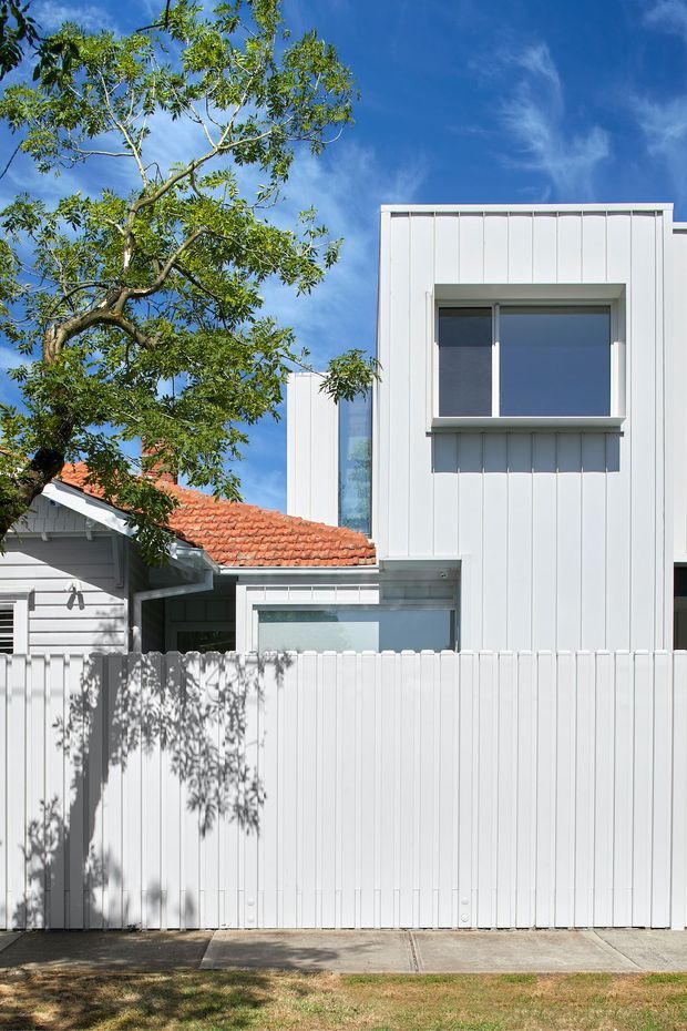 Exterior view of a contemporary residential extension behind a tall white vertical-panel fence. The composition combines a modern white-clad two-storey form with large rectangular windows and a lower glazed connection, alongside an older weatherboard structure with a terracotta tiled pitched roof. A leafy tree partially frames the facade under a bright blue sky.