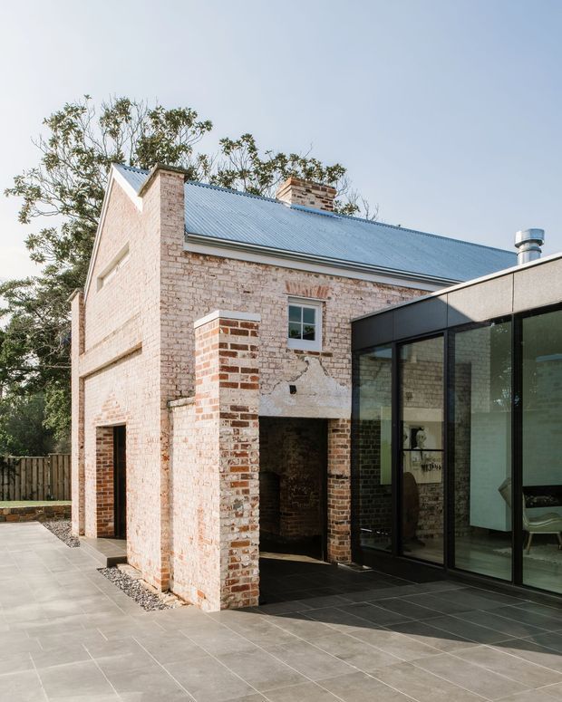 Exterior view of a renovated brick building with a pitched corrugated metal roof and prominent masonry chimneys. A contemporary black-framed glazed extension adjoins the original structure, opening onto a large tiled terrace. The image contrasts historic brick fabric with modern minimalist glazing and dark paving.