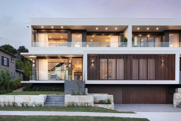 Contemporary multi-storey residence viewed from the street at dusk, featuring crisp white rendered volumes, extensive floor-to-ceiling glazing, glass balustrades, recessed warm lighting, vertical timber screening, a covered entry stair, landscaped frontage, and an integrated double garage.