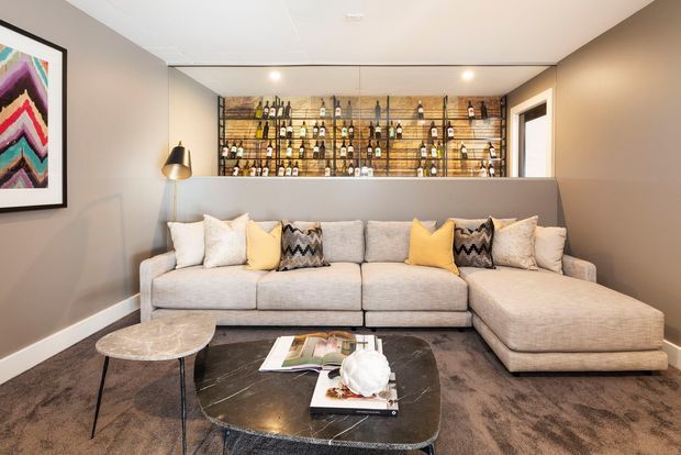 A stylish living room with a large light-grey L-shaped sectional sofa, patterned and yellow accent cushions, dark marble coffee tables, and a deep-toned carpet. A timber-backed built-in shelving and display area with bottles forms a feature wall behind the sofa, complemented by warm integrated lighting and neutral painted walls.