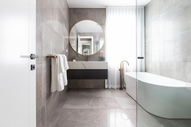 A refined contemporary bathroom with large-format grey stone-look wall and floor tiles, a freestanding white bathtub, frameless glass shower enclosure, floating dark vanity, round brass-framed mirror, and a bright sheer-curtained window. White towels and warm brass plumbing accents add contrast to the cool neutral palette.