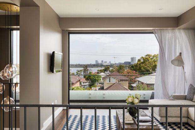 Contemporary upper-level living space with a large framed window overlooking a suburban waterfront and city skyline. The room features a neutral palette, sheer curtains, a grey sofa, wall-mounted television, sculptural pendant lighting, black metal balustrade, timber flooring, and a bold blue-and-white geometric rug.