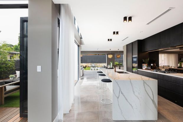 A bright, elongated contemporary kitchen with a large white veined stone island, black built-in cabinetry, integrated appliances, and minimalist pendant-style ceiling lighting. Floor-to-ceiling glazing and sliding doors connect the kitchen to an outdoor area, while bar stools line the island.