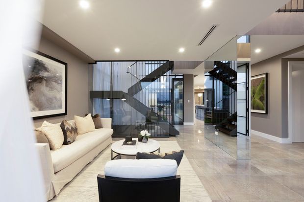 A refined contemporary living room opening into a dramatic double-height stair hall. The space features a cream sectional sofa, dark lounge chair, round coffee table, pale rug, polished light stone flooring, grey walls, extensive glass balustrades and internal glazing, and a sculptural dark stairway with black metal detailing. Large glazing brings in abundant natural light, while mirrored surfaces visually expand the room.