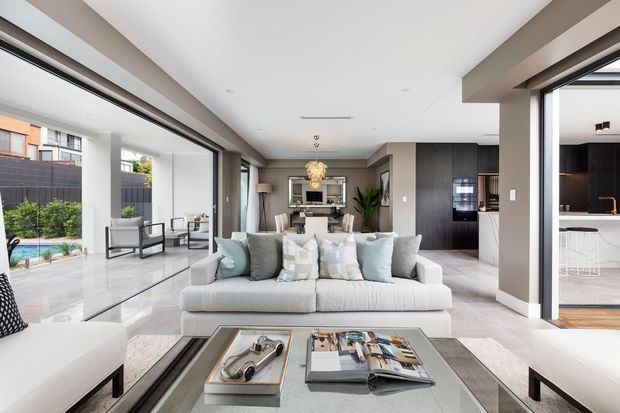 Bright contemporary open-plan living area with a large light-grey sectional sofa facing a glass coffee table. Floor-to-ceiling glazing opens to a patio and pool area, while the rear of the room connects to a dining area and a sleek kitchen with dark cabinetry and a white island. Neutral finishes, recessed lighting, and a feature pendant create a refined, airy interior.