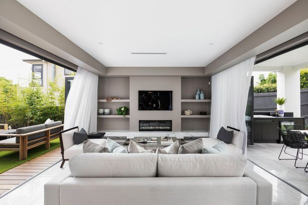 Bright contemporary open-plan living room with a large white sectional sofa facing a built-in media wall and fireplace. Floor-to-ceiling glazing and sheer curtains open onto a landscaped outdoor area and a covered outdoor kitchen or entertaining zone. The room features recessed lighting, grey built-in shelving, pale flooring, and a neutral white, grey, and timber palette.