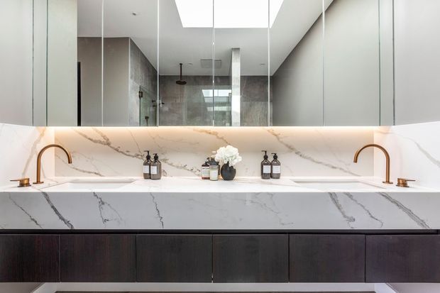 Contemporary bathroom vanity with a long white marble-look benchtop and matching full-height splashback, twin curved brass faucets and undermount basins. Dark timber cabinetry forms the vanity base, while a large mirrored wall reflects a tiled shower area and skylight above.