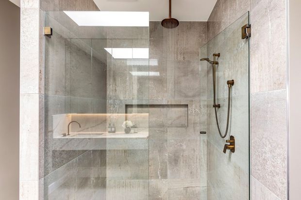 Contemporary walk-in shower enclosed by clear frameless glass panels, with large-format grey stone-look wall tiles, a recessed niche, and bronze shower fixtures. A floating vanity with a vessel basin is visible through the glass reflection.