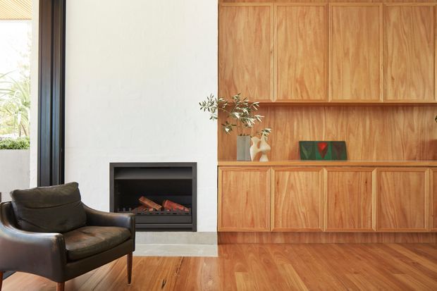 Warm contemporary living room featuring a built-in timber joinery wall with upper and lower cupboards, open display shelving, and a dark built-in fireplace set into a pale plaster wall. A dark leather lounge chair sits beside the fireplace, while timber flooring and a large black-framed glazed opening contribute to the inviting, refined interior.