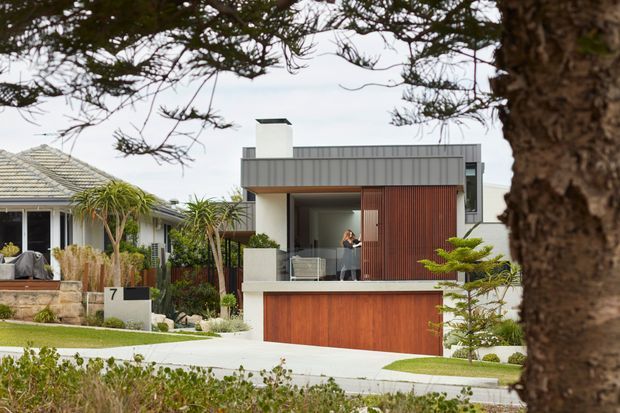 Street-facing view of a contemporary multi-level residence with a flat roof, strong horizontal forms, grey metal-like fascia, warm vertical timber-clad screening, and a timber garage door. The home is set within a landscaped suburban streetscape, with mature trees and neighbouring residences visible.