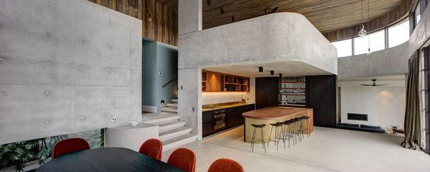 A dramatic open-plan interior with a concrete sculptural mezzanine or upper volume, curved timber ceiling, dark timber kitchen cabinetry, and a rounded timber island with stools. The space includes a dining table with red chairs in the foreground, a stepped circulation area, and a sunken or lower living zone with expansive curved glazing. The palette is warm timber, raw concrete, dark joinery, and muted neutral finishes.
