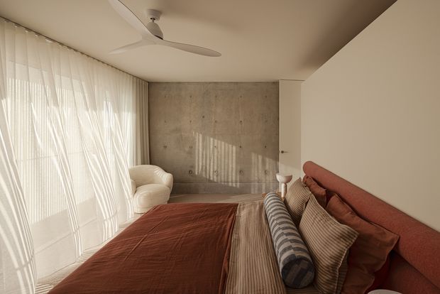 Warm, softly lit contemporary bedroom with a large upholstered bed, layered neutral and rust-toned bedding, a cream armchair, sheer floor-to-ceiling curtains, a minimalist ceiling fan, and a raw concrete feature wall. Natural light filters through the curtains, creating a calm, refined atmosphere.
