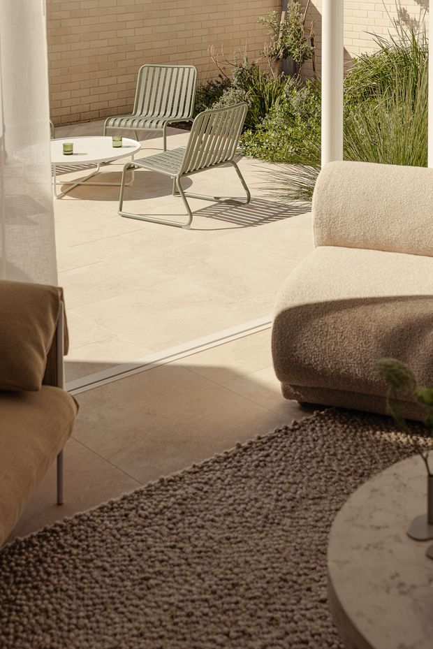 A warm, sunlit living room opens directly onto a paved outdoor terrace through wide glazed sliding doors. The interior features a large textured neutral rug, rounded upholstered lounge seating, and a pale stone or terrazzo coffee table. Outside, two modern slatted metal lounge chairs and a low white table sit against a light tan brick wall, with dense ornamental planting along the edge.